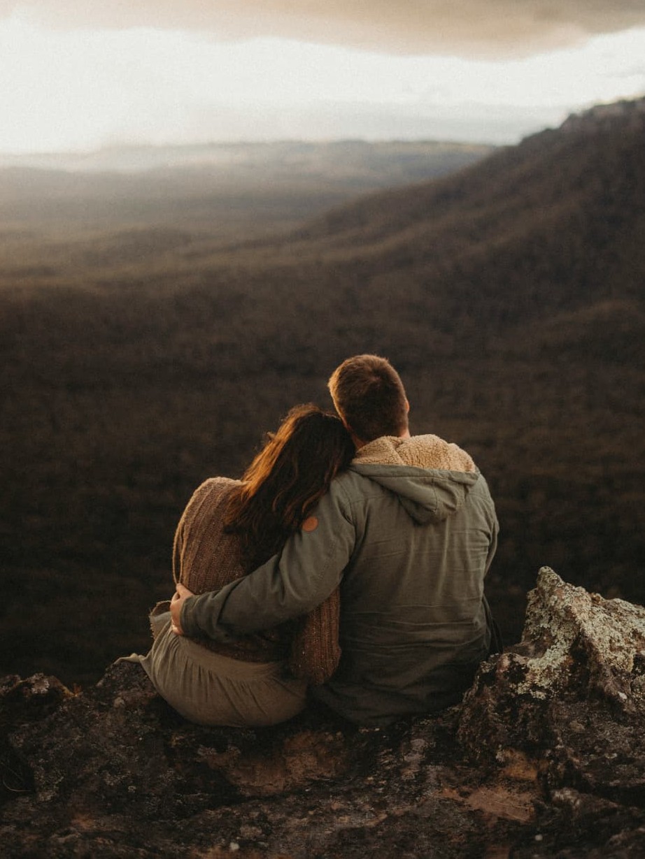 A couple sitting close together on a clifftop, looking out over a forested valley at dusk